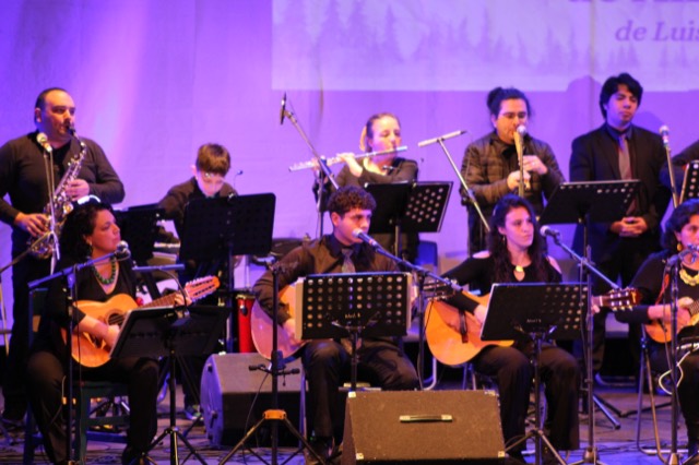 Integrantes de Ensamble Clase con guitarras, charangos, clarinete y flauta traversa, iluminados en tonos violeta.