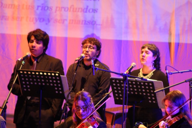 Tres cantantes de Ensamble Clase frente a sus micrófonos, con violines en primer plano.